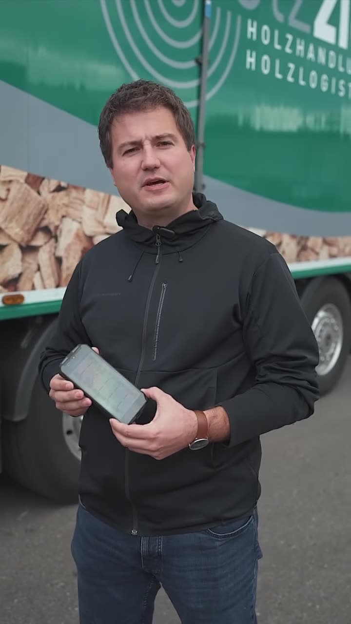 Mitarbeiter zeigt die Reifendruck-App auf dem Smartphone vor einem beladenen Holztransporter